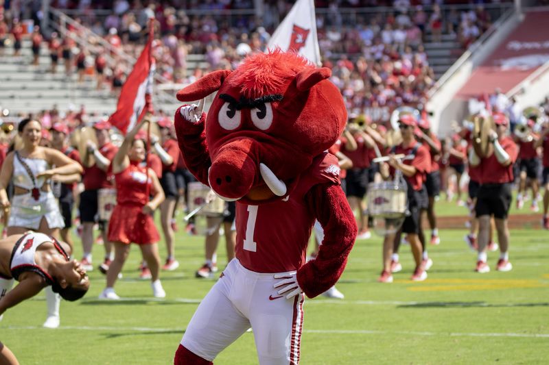 A red hog mascot poses on the field in front of cheerleaders and the band