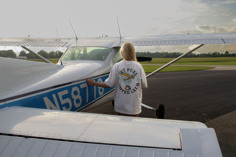 A woman in a Jet Fuel Coffee Club tee leans on a small plane