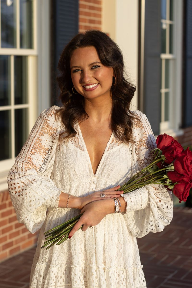 A young woman in white lace holds red roses outside a brick building