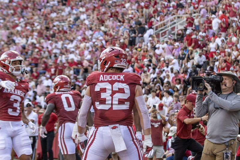 Arkansas football players, number 32 in front, walk onto the field past a TV camera operator
